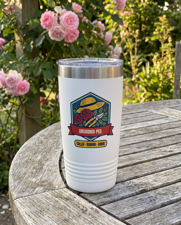 Rose Garden Badge, a rose garden badge in teal, red, gold, green and navy, UV printed on the front of a 20oz Ringneck tumbler in White, sample name GRANDMA PEG, sits on a garden patio table, climbing pink roses far behind in summer sun