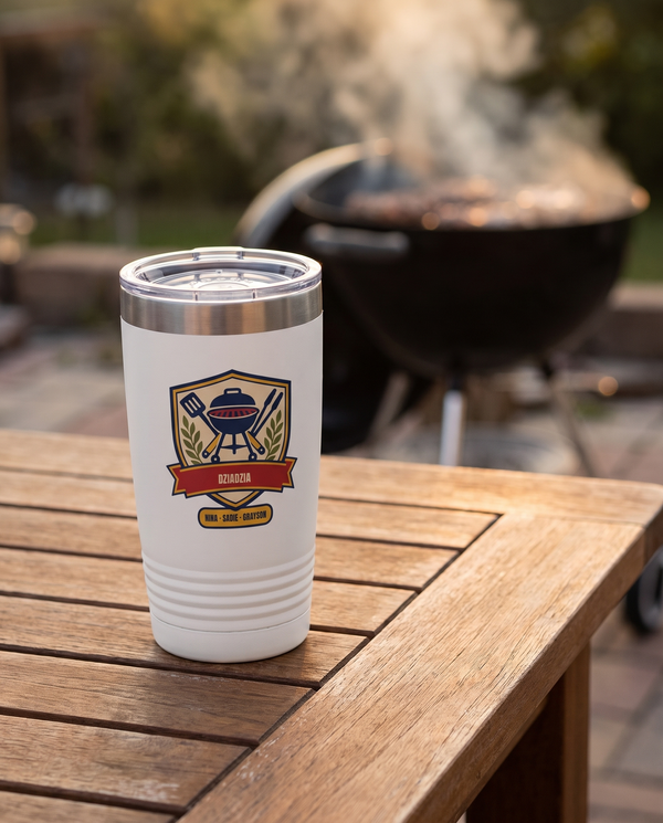 Backyard Grill Badge, a grill badge in navy, red, gold, cream and olive, UV printed on the front of a 20oz Ringneck tumbler in White, sample name DZIADZIA, sits on a wide wooden patio table whose top fills the whole lower part of the frame edge to edge, a charcoal grill with soft smoke far behind, evening