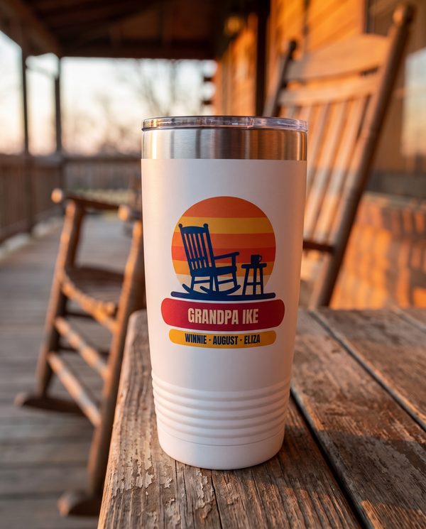 Rocking Chair Sunset, a porch sunset in navy, orange, gold and red, UV printed on the front of a 20oz Ringneck tumbler in White, sample name GRANDPA IKE, sits on a porch table at sunset, a wooden rocking chair far behind, orange sky