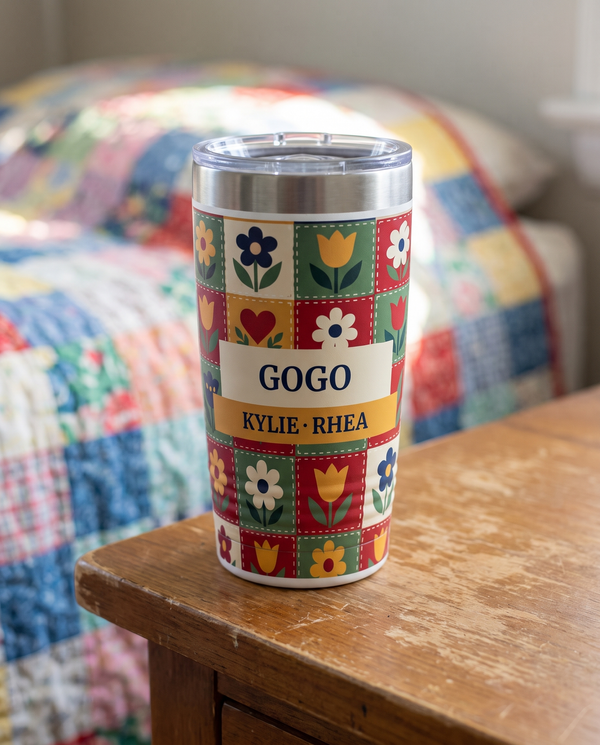 Patchwork Quilt, a folk patchwork quilt in red, green, gold, cream and navy, printed all the way around a 20oz Ringneck tumbler in White, sample name GOGO, sits on a wooden nightstand, a patchwork quilt on a bed far behind, soft morning light