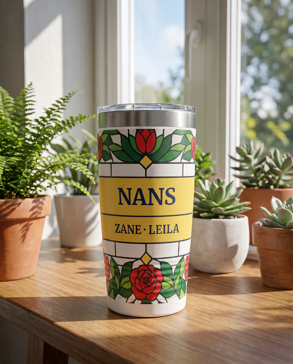 Stained Glass Garden, stained glass roses, tulips and irises in red, blue, green and gold, printed all the way around a 20oz Ringneck tumbler in White, sample name NANS, sits on a sunroom table with potted plants and a tall window far behind, bright day