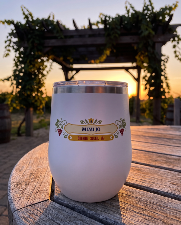 Folk Grapevine, a folk grapevine in red, sage, gold and cream, UV printed on the front of a 12oz stemless wine tumbler in White, sample name MIMI JO, sits on a patio table at sunset, grapevines on a pergola far behind