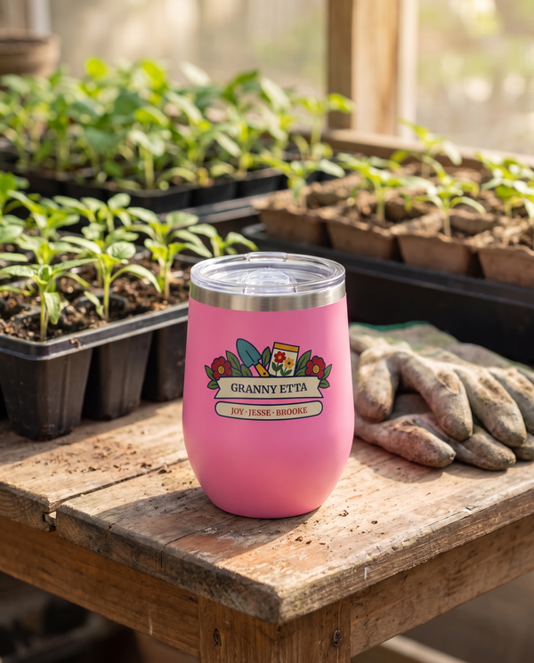 Seed Packet Garden, a garden scene in red, teal, gold, green and cream, outlined in navy, UV printed on the front of a 12oz stemless wine tumbler in Pink, sample name GRANNY ETTA, sits on a potting table with trays of seedlings and garden gloves far behind, spring light