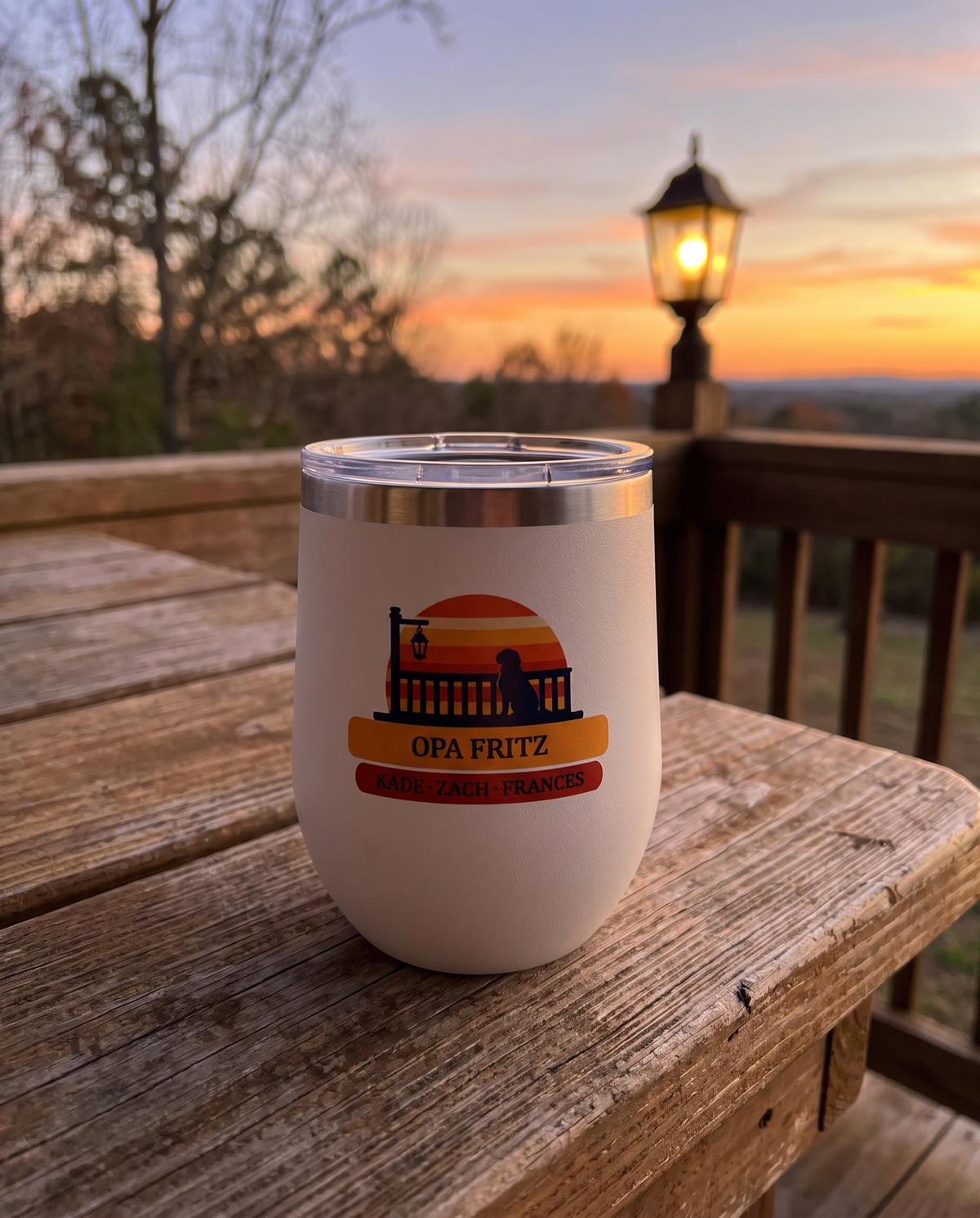 Porch Dog Sunset, a porch sunset with a dog in navy, orange, gold and rust, UV printed on the front of a 12oz stemless wine tumbler in White, sample name OPA FRITZ, sits on a porch railing table at sunset, a lantern on a post far behind