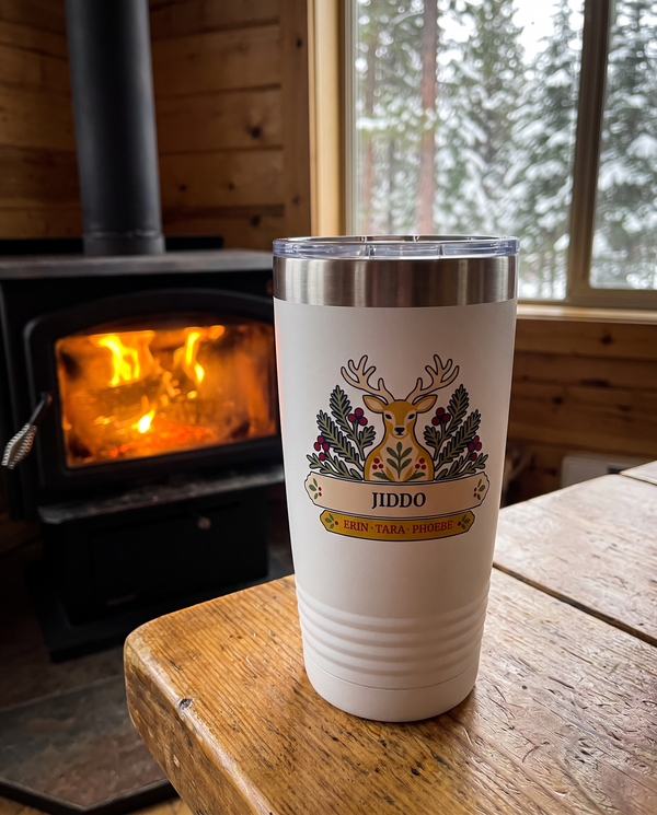 Folk Deer, a folk deer and pine boughs in gold, cream, red, navy and sage, UV printed on the front of a 20oz Ringneck tumbler in White, sample name JIDDO, sits on a cabin table near a wood stove glow, snowy pines through a window far behind