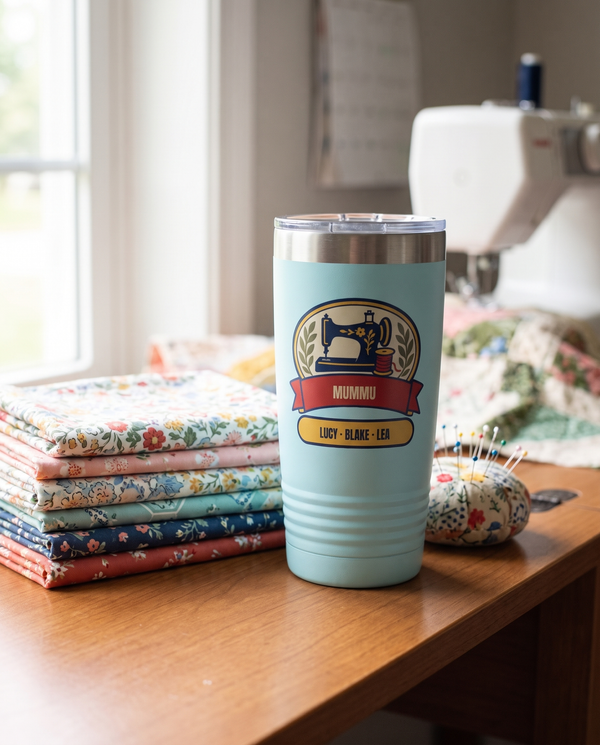 Sewing Machine Badge, a sewing badge in navy, red, cream, gold and olive, UV printed on the front of a 20oz Ringneck tumbler in Light Blue, sample name MUMMU, sits on a sewing table beside folded quilting fabric and a pincushion, soft window light