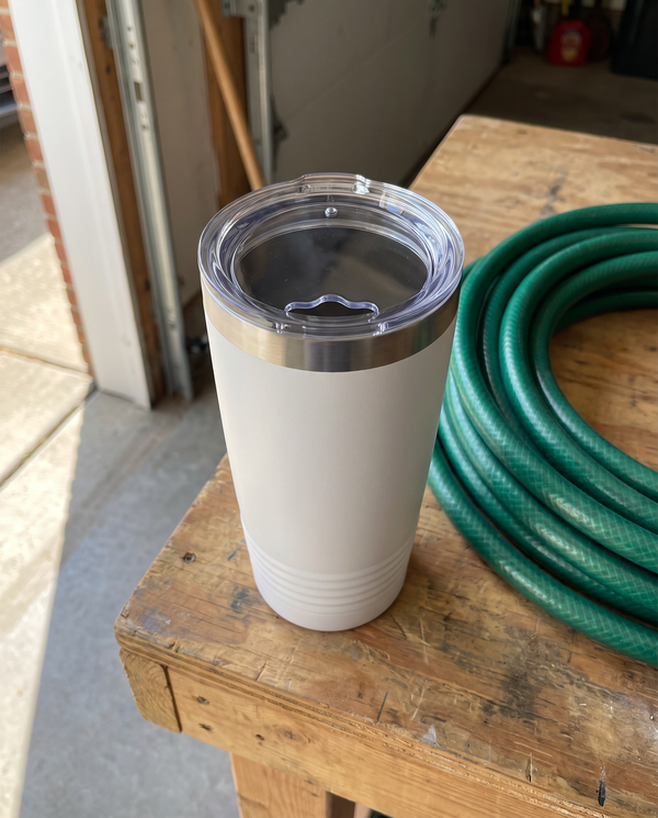 Clear standard lid with its open sip hole on a white 20oz Ringneck tumbler, garage workbench by a coiled hose